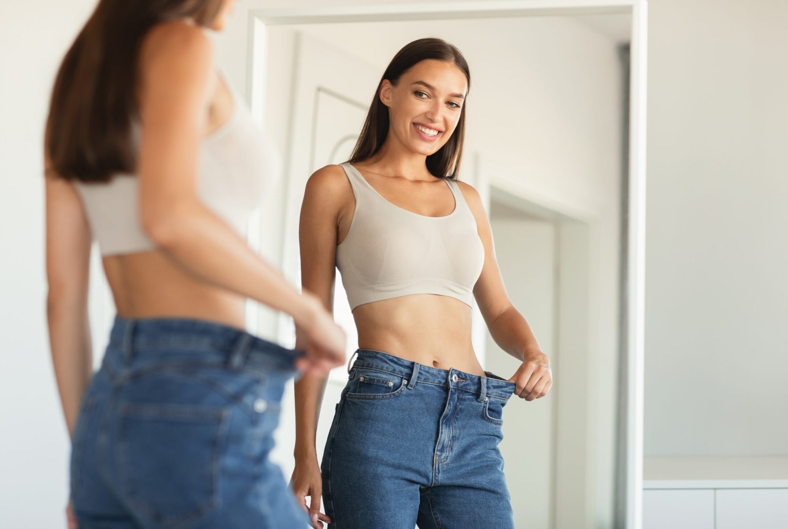 A happy woman holds out loose jeans in front of a mirror, showing weight loss success after body contouring at a Hayden Idaho med spa. at North Idaho Med Spa in Hayden